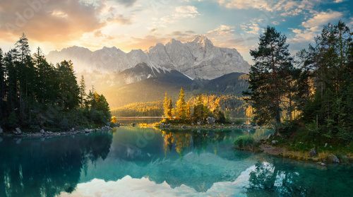 Painterly lake scenery in Germany with mountains reflected in the teal water and a beautiful ray of morning sunlight illuminating some trees